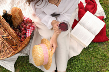 Woman with glass of wine, book and picnic basket on green grass, above view