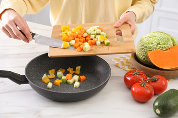 Woman pouring mix of cut vegetables into frying pan at table in kitchen, closeup - obrazy, fototapety, plakaty