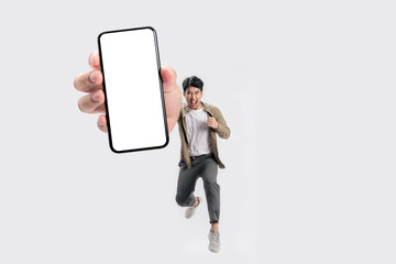 Full length of cheerfull Asian man jumping and smiling in air with showing cellphone blank screen...