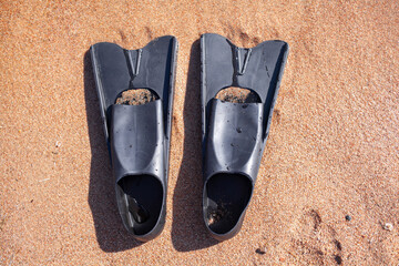 A pair of black flippers on the background of sand next to the water, top view. Swimming equipment - fins on the shore. Summer holidays, fun, exploring the sea world concept. Space for copy.