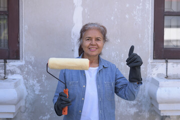 Front view medium shot portrait of happy senior Asian woman wearing gloves, holding paint roller, smiling, putting thumb up. Worker, retiree lifestyle, DIY home renovation concept.