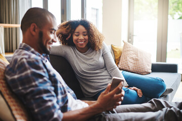 Happy, carefree and relaxed couple reading text on a phone while sitting on a couch. Man and woman smiling and laughing while browsing an online social media app or watching funny videos at home
