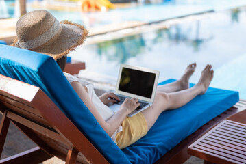 Unrecognizable woman sitting or lying down on blue outdoor pool chair, using laptop computer,...