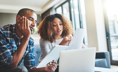 Black couple talking, reading finance papers and planning their bills while working on tablet and laptop on the couch together at home. Husband and wife going through paperwork for tax and loans