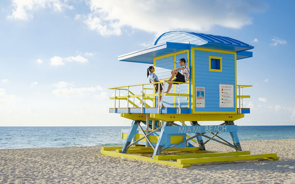 Miami Beach, a couple on the beach in Miami Florida, lifeguard hut Miami Asian women and caucasian men on the beach during sunset. man and woman relaxing at a lifeguard hut looking at ocean - Powered by Adobe