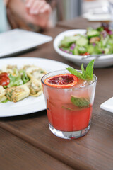 A view of a red colored mint and blood orange garnished cocktail, among several assorted restaurant entrees.
