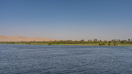 There are ripples on the surface of the blue wide river. On the shore there is green vegetation, palm trees. Sand dunes against a clear sky. Egypt. Nile. Copy Space