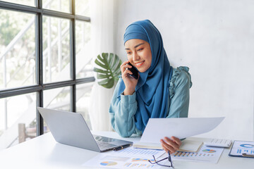 Successful pretty Asian muslim lady in hijab make a phone call when working at desk in office during working on her project on laptop computer.