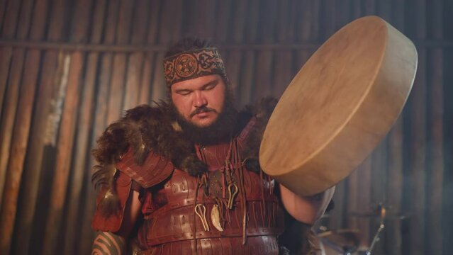 Man in spiritual ecstasy with closed eyes taps shamanic drum with special furry stick doing ritual closeup. Cosplayer in Altai national suit plays on stage