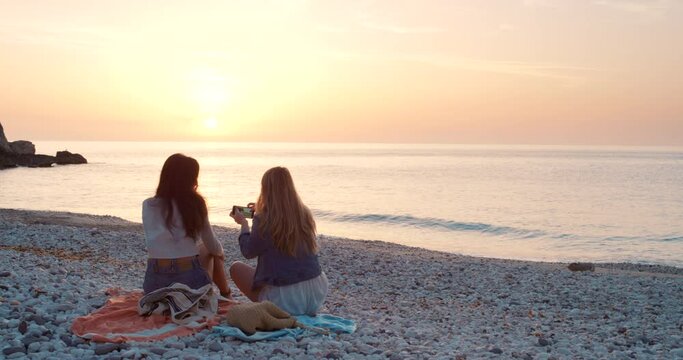 Relaxed friends taking a photo with a phone while enjoying the view at the sunset beach. Rear view of women on vacation on a tropical island during summer and posting with an online social media app