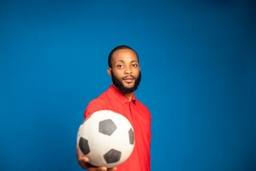 young African Nigerian man in casual red shirt posing isolated on blue background studio portrait. People lifestyle concept. holding football showing in to the camera