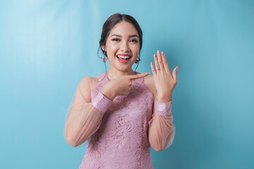 Smiling young Asian woman wearing modern kebaya showing her engagement ring isolated over blue...
