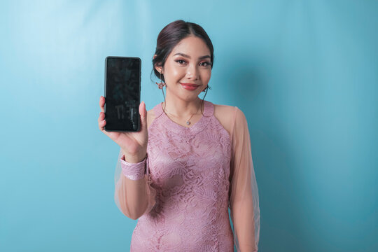 A portrait of a cheerful Asian woman showing copy space on her smartphone over blue background, wearing a modern kebaya