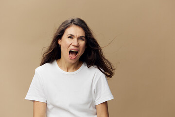 a beautiful, happy woman standing on a beige background with long hair developing in the wind, standing in a white T-shirt screaming loudly