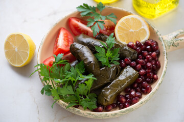 Middle close-up of dolma or dolmades with pomegranate seeds, tomato wedges, fresh parsley and lemon, horizontal shot on a light-beige marble background