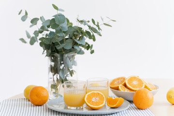 two glasses of orange juice, fresh oranges and lemons and eucalyptus branches in glass vase on wooden table in white kitchen