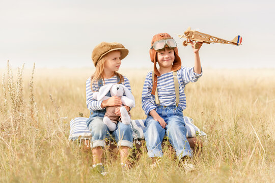 Boy and a girl are sitting on a suitcase in the grass under the open sky. Cheerful and happy children play in the field and imagine themselves to be pilots on a sunny summer day. Children dream of fly