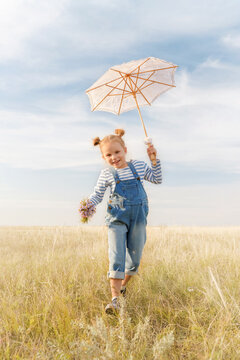 Child plays in a field on a summer day. Little girl with a bouquet of flowers and an umbrella against the blue sky. Cheerful and happy kid dreams of flying.