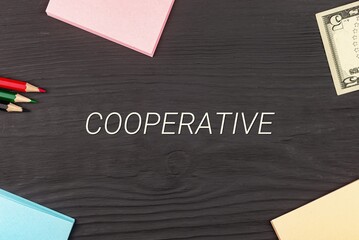 COOPERATIVE - text, money dollars, stickers and colored pencils on a black wooden table. Business concept: buying, selling, commerce (copy space).
