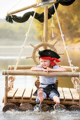 Child in a pirate costume plays on a wooden raft at sunset. Girl pretends to be the captain of a...