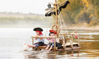 Children in pirate costumes play on a wooden raft at sunset. Two girls pretend to be the captains of a ship with black sails and a flag. Funny kids dreaming about adventure and travel. - 521603984