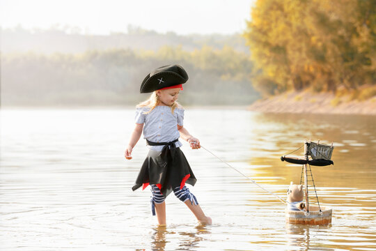 Child in a pirate costume plays with a toy boat at sunset. Girl pretends to be the captain of a ship with black sails and a flag. Funny child dreams of adventure and travel.