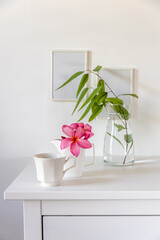 Plumberia rumba in a glass, eucalyptus branch in a vase and cup of coffee on a white table against a wall with two frames. Ready layout. Vertical frame. Space for text