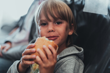 little candid kid boy five years old eats burger or sandwich food sitting in airplane seat on flight traveling from airport. children take a bite. child in air plane eating lunch or dinner meal