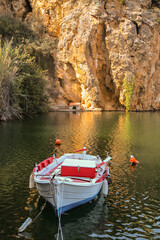 A fishing boat is anchored on Lake Voulismeni, next to the rock that surrounds the lake. The boat and the rocks colored by the sunset light are reflected in the clear water.