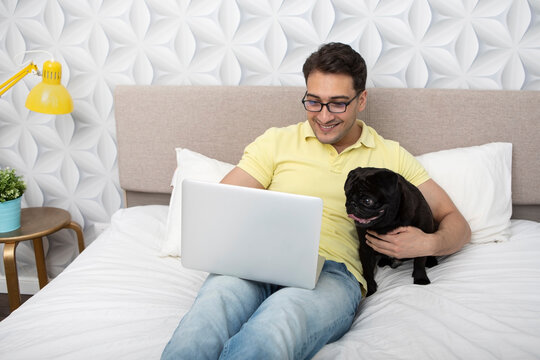 Smiling happy man sitting on the bed and hugging his lovely pet black pug breed and working on laptop. Friendship concept