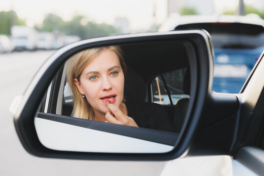 Beautiful woman with blond hair driving a car paints her lips with lipstick. The woman driving making make-up. Reflection of a woman in the rearview mirror. Photo of a successful woman in a car.