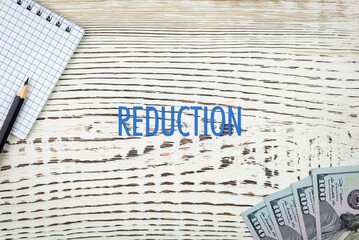 Naklejka premium REDUCTION - word (text) and dollars on a white wooden table, notebook, notepad. Business concept: buying, selling, commerce (copy space).