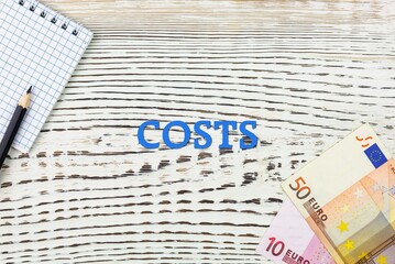 Naklejka premium COSTS - word (text) and euro money on a white wooden table, notebook, notepad. Business concept: buying, selling, commerce (copy space).