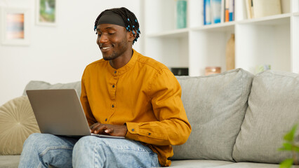 Smiling black man using computer working or studying while sitting on sofa at home. Free copy space