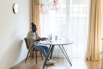 Happy young black man using laptop computer for online work at table in home office, . Cool African American guy having remote job, freelancing on web. Modern business concept