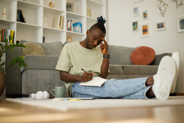 Pensive black teenager writing, taking notes, studying in a notebook while sitting on the floor at home