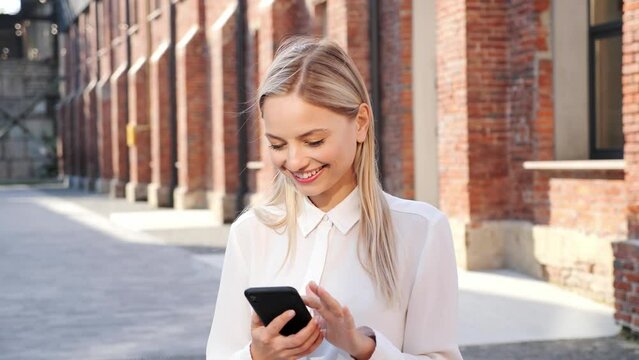Happily Smiling Attractive Woman Standing on the Street. Typing on her Smartphone with Interest. Enjoying Free time Scrolling Social Media using Internet Data App. Beautiful Girl Posting New Photos.
