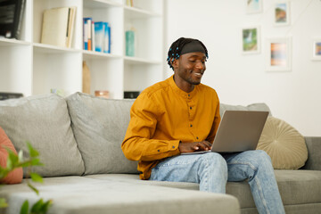 Black african man in a shirt with a laptop sits on a sofa in the living room, free copy space.