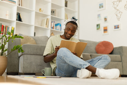  Smart black teenage student in full growth with a book makes notes. Smart teenager writing assignment or term paper, studying remotely indoors