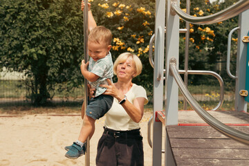 Grandmother supporting and holding her 2 years old grandson at children playground. Grandmother and child having fun. Support childhood symbol. Sunny, summer day