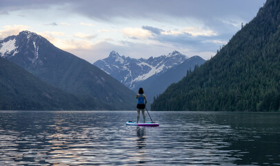 Fototapeta na wymiar Adventurous Woman Paddle Boarding in a Lake around Canadian Mountain Landscape. Chilliwack Lake, British Columbia, Canada. Adventure Sport Travel
