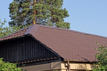 attic of a wooden rural house under a brown tiled roof on the street against the background of a blue sky and a green pine tree