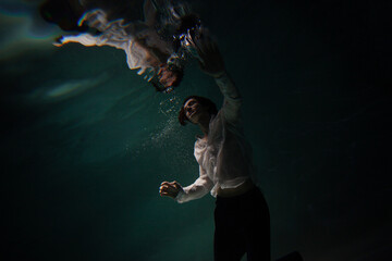 Obraz na płótnie Canvas Photo underwater, a guy in a white shirt floundering underwater and reaching for the surface of the water, a man and his reflection. mystical underwater portrait