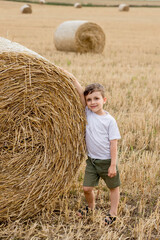 Naklejka na ściany i meble A cute little boy in a white t-shirt and khaki shorts is standing near round bales of hay. Photo session in the field