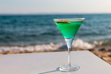 Fresh cold green cocktail in a martini glass with lemon on the beach with sea in background.Summer sea vacation and travel concept. Copy space