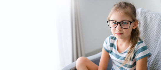 Child girl in glasses at home. Portrait of beautiful blonde teenager sitting on sofa. Charming...