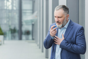 Senior male businessman sick near office, boss uses inhaler to ease breathing, gray haired banker has asthma attack