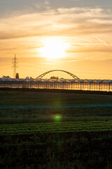 Fototapeta premium Golden sunset over greenhouse silhouettes with bridge and electricity tower for solar power in agricultural business on idyllic countryside and rural scenery shows glass greenhouses healthy vegetables