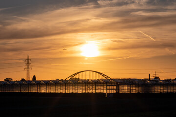 Golden sunset over greenhouse silhouettes with bridge and electricity tower for solar power in agricultural business on idyllic countryside and rural scenery shows glass greenhouses healthy vegetables