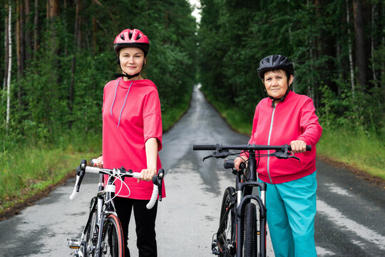 Mother, daughter with bicycles in sportive helmets, suits look at camera, standing on road surrounded by trees. Young sports coach and senior woman are going to ride bicycles in forest
